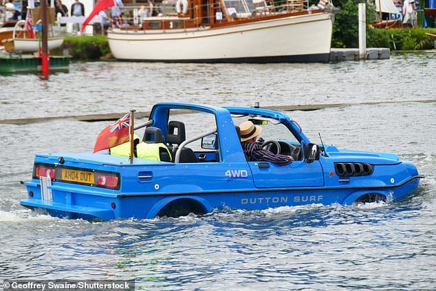Автомобиль-амфибия 4WD Dutton Surf движется по воде, за рулем сидит водитель в стильном полосатом пиджаке.